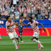 United States forward Folarin Balogun celebrates after scoring against Paraguay during a World Cup match as teammate Weston McKennie runs toward him and fans erupt in the stands.