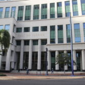 Exterior view of the San Diego Hall of Justice in downtown San Diego, a multi-story courthouse building with tall white columns, large windows, and pedestrians walking along the sidewalk.