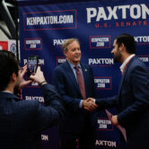 Men in suits shaking hands while another man photographs them on a cell phone.